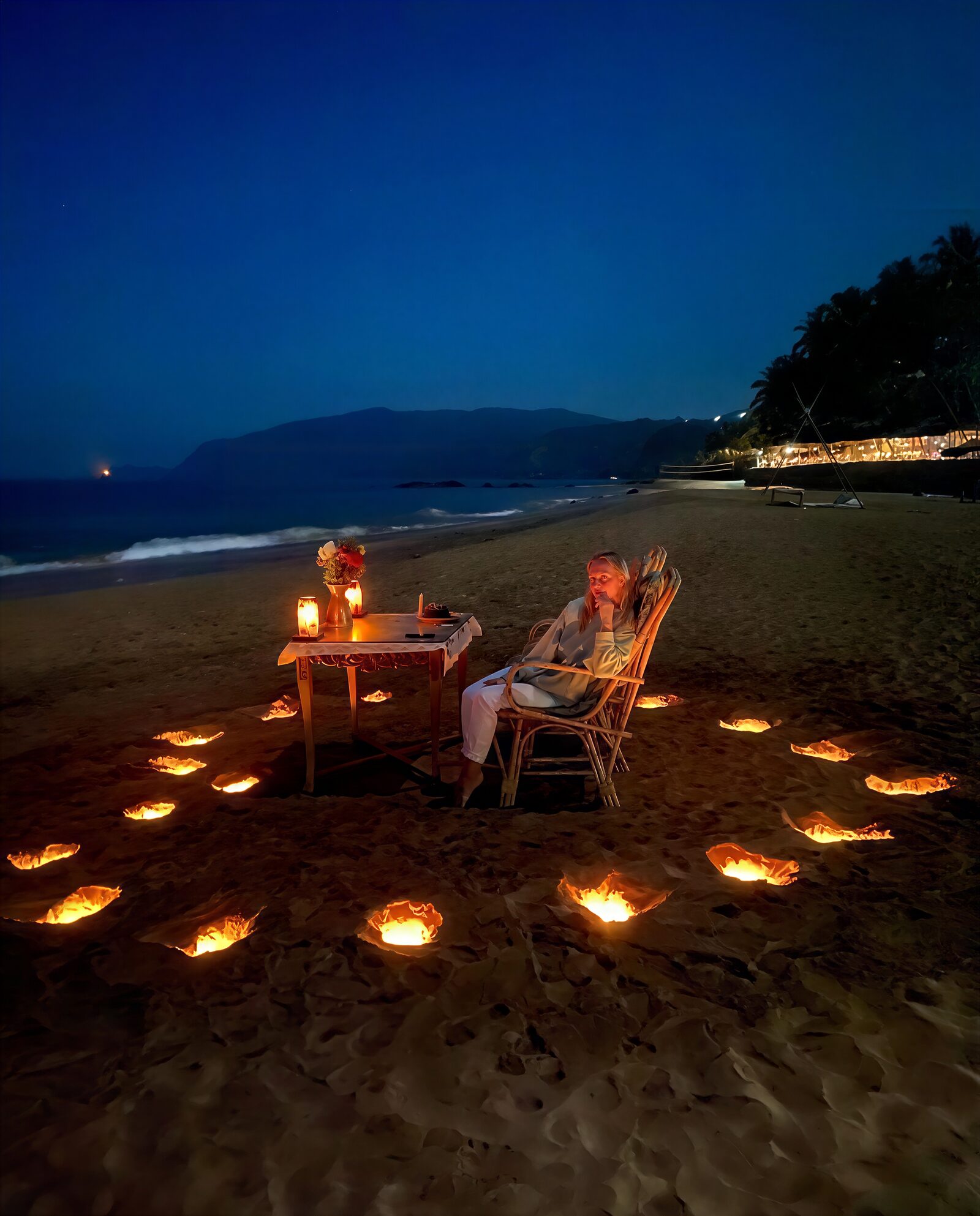 A candlelit table for two on the sand at dusk, ringed with lanterns